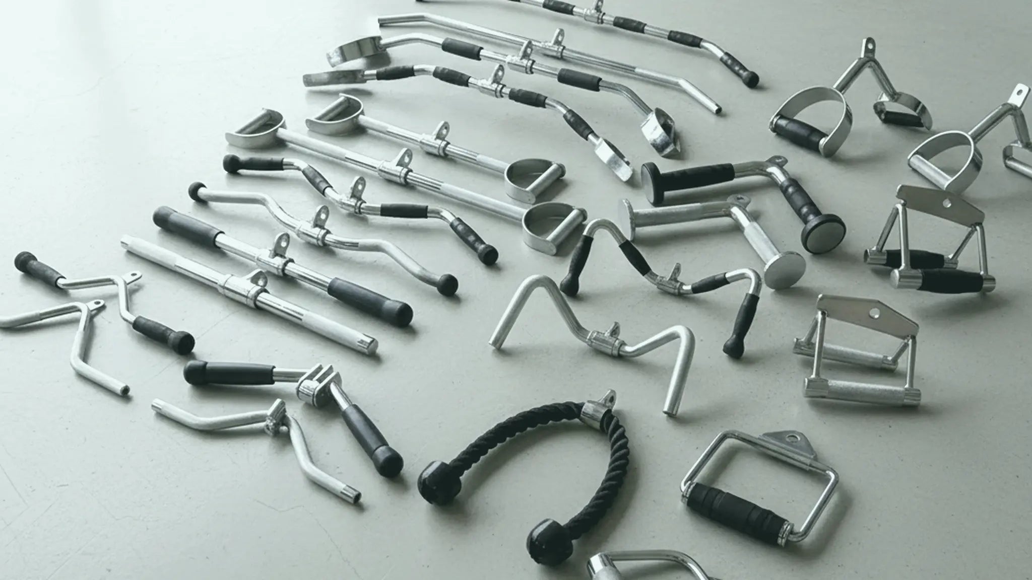 Comprehensive array of silver and black cable machine attachments, including tricep ropes, lat bars, V-handles, and D-handles, displayed against a dark, seamless background.