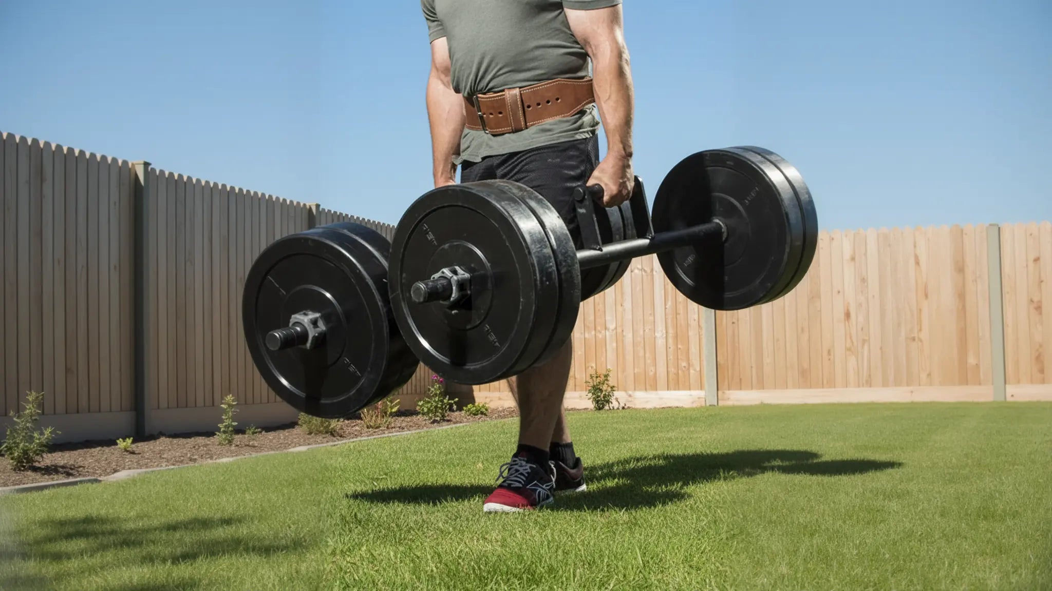 Man performing a farmer's walk exercise in a home garden with minimal plants, holding heavy farmers walk handles at his sides.