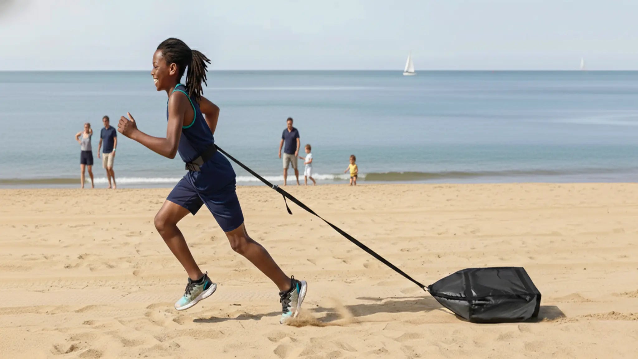 Young boy in fitness attire pulling a black drag sled on a sandy beach with the sea in the background, demonstrating fun, low-impact training for all ages.