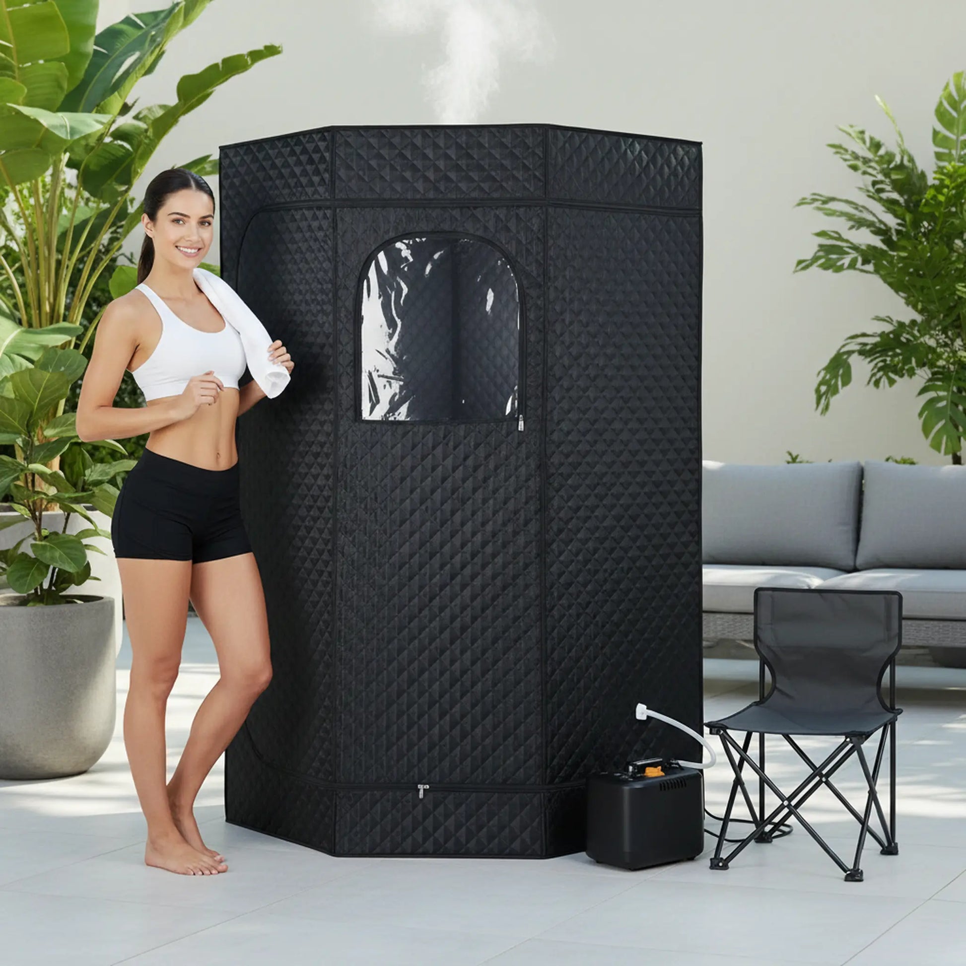 Woman standing next to a black portable sauna in an outdoor setting with plants and a couch.