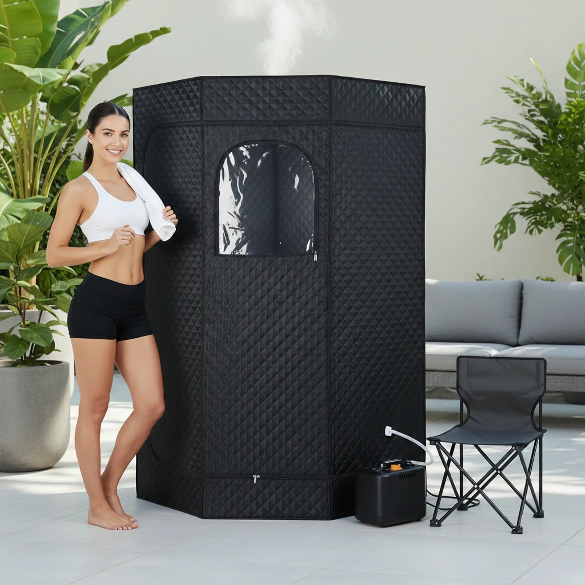 Woman standing next to a black portable sauna in an outdoor setting with plants and a couch.