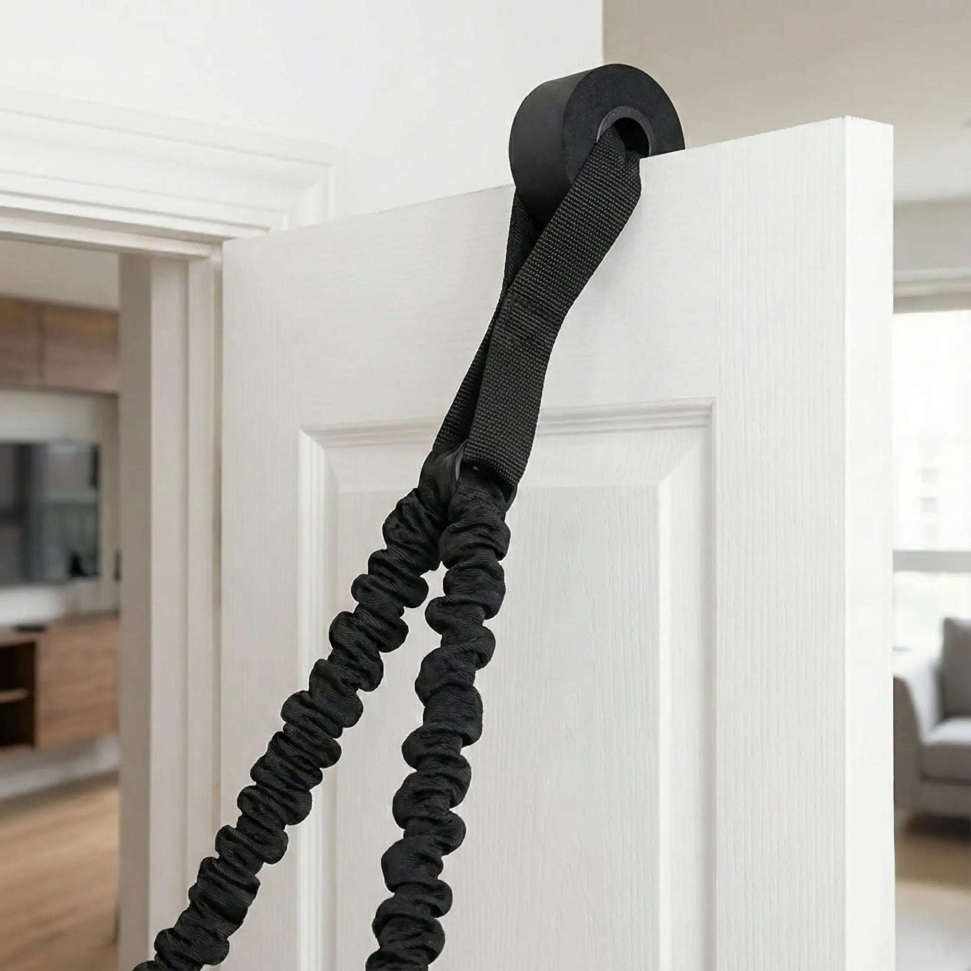 Close-up view of a resistance band door anchor strap securely fastened in a white door frame, with the heavy-duty black nylon band threaded through for home training.
